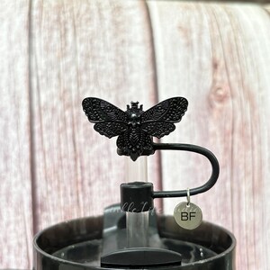 May include: Black water bottle with a decorative butterfly-shaped stopper. The stopper is detailed with intricate wing patterns and sits atop a black handle. A small, round charm with the letters "BF" hangs from the handle.