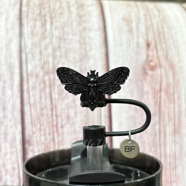 May include: Black water bottle with a decorative butterfly-shaped stopper. The stopper is detailed with intricate wing patterns and sits atop a black handle. A small, round charm with the letters "BF" hangs from the handle.