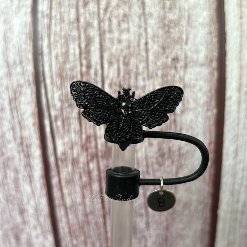 May include: A black, decorative butterfly-shaped straw topper. The topper is made of metal and features intricate details. A small, round charm with the letter "B" hangs from the straw holder. The straw is clear and the background is a wooden surface.