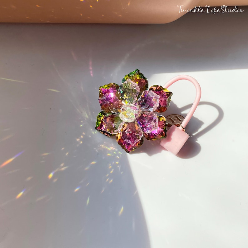 May include: A pink hair tie with a multi-colored crystal flower charm. The flower has green, pink, and clear crystals.
