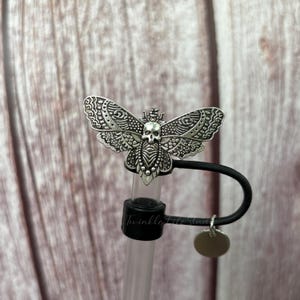 May include: A silver-colored moth-shaped bottle stopper with a skull design. The moth has detailed wing patterns and is attached to a black rubber stopper. A small, round silver charm hangs from the stopper.