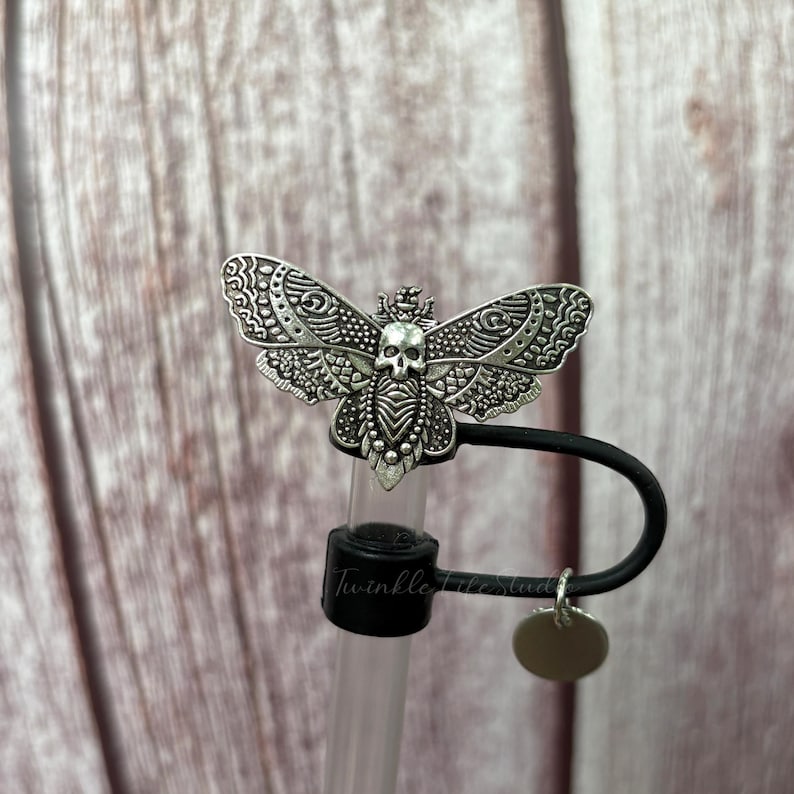 May include: A silver-colored moth-shaped bottle stopper with a skull design. The moth has detailed wing patterns and is attached to a black rubber stopper. A small, round silver charm hangs from the stopper.