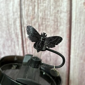 May include: A black metal butterfly-shaped straw topper. The butterfly has intricate wing details and a small crown. The straw topper is attached to a black lid with a clear straw. The text "Twinkle Life Studio" is visible.