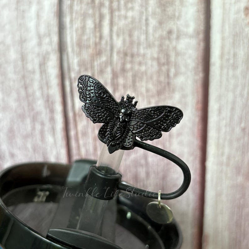 May include: A black metal butterfly-shaped straw topper. The butterfly has intricate wing details and a small crown. The straw topper is attached to a black lid with a clear straw. The text "Twinkle Life Studio" is visible.