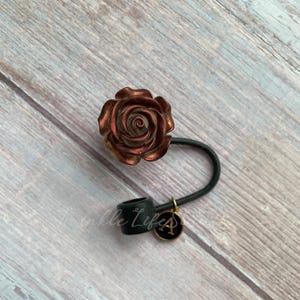 Pode incluir: Uma fivela de cabelo em forma de rosa em tons de cobre e castanho. A fivela de cabelo tem uma faixa preta e um pequeno pingente com a letra "A". O texto "Twinkle Life" &eacute; vis&iacute;vel na base preta.