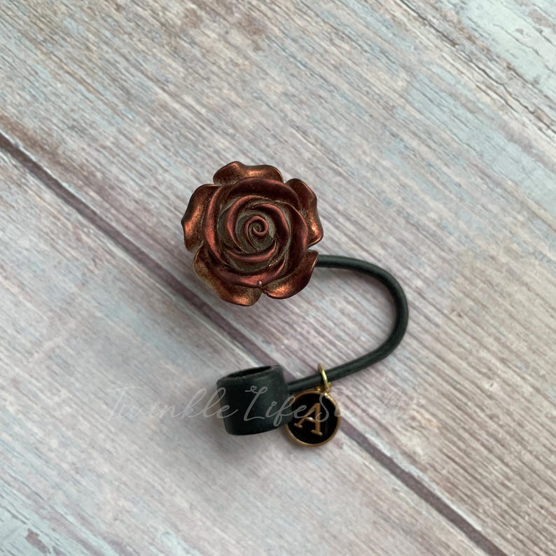 Pode incluir: Uma fivela de cabelo em forma de rosa em tons de cobre e castanho. A fivela de cabelo tem uma faixa preta e um pequeno pingente com a letra "A". O texto "Twinkle Life" &eacute; vis&iacute;vel na base preta.