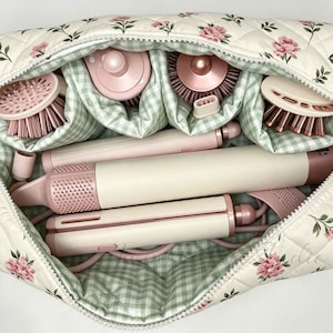 Op de afbeelding: Een gewatteerde, crèmekleurige reisetui met een roze bloemenpatroon en een lichtgroene gingham voering. De etui bevat een haarstyling-set, inclusief een handvat, een stijltang en vier verschillende ronde borstelopzetstukken.