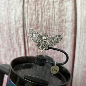 May include: A silver-colored moth-shaped bottle stopper with a skull design. The stopper is attached to a black handle and a black lid. The text "Twinkle life" is visible on the clear plastic part of the stopper. A small silver charm with the letters "BR" hangs from the handle.