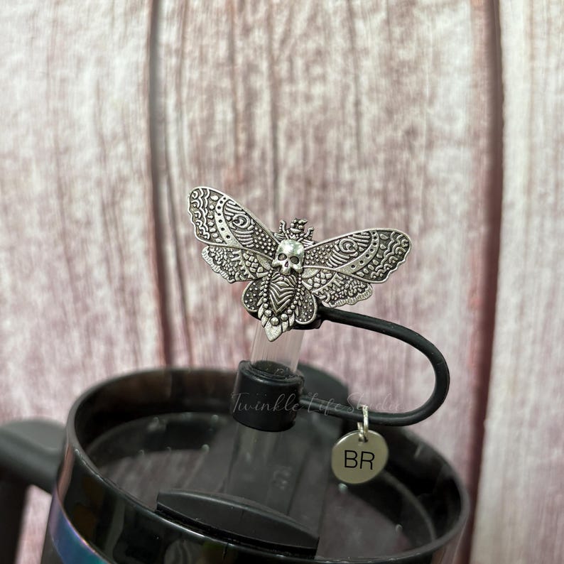 May include: A silver-colored moth-shaped bottle stopper with a skull design. The stopper is attached to a black handle and a black lid. The text "Twinkle life" is visible on the clear plastic part of the stopper. A small silver charm with the letters "BR" hangs from the handle.