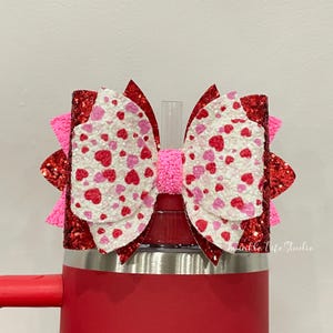 May include: A red glitter bow with a white layer covered in red and pink hearts, atop a red tumbler. The bow has a pink center and pink accents. The tumbler has a silver rim and a red handle.