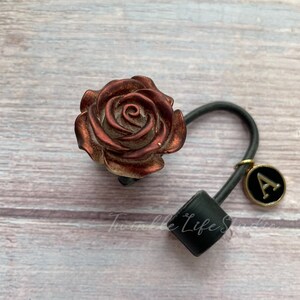 Pode incluir: Uma fivela de cabelo em forma de rosa em tons de bronze e cobre. O acess&oacute;rio de cabelo apresenta uma faixa el&aacute;stica preta e um pequeno pingente com a letra "A" em um fundo preto. As palavras "Twinkle Life Studio" s&atilde;o vis&iacute;veis.