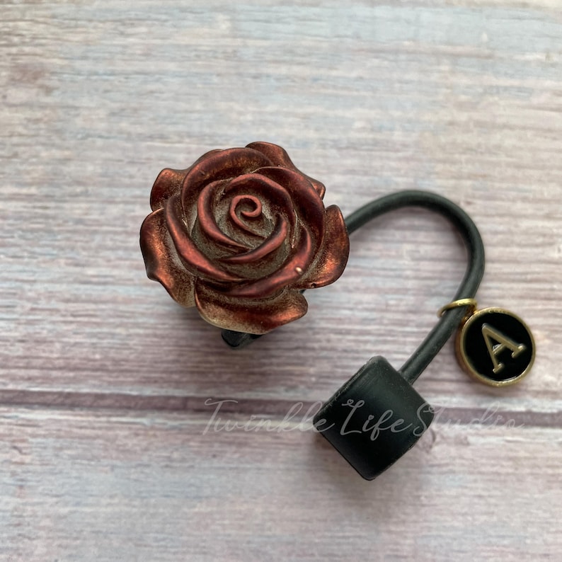 Pode incluir: Uma fivela de cabelo em forma de rosa em tons de bronze e cobre. O acess&oacute;rio de cabelo apresenta uma faixa el&aacute;stica preta e um pequeno pingente com a letra "A" em um fundo preto. As palavras "Twinkle Life Studio" s&atilde;o vis&iacute;veis.