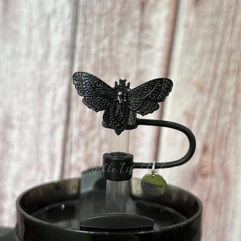 May include: Black bottle stopper featuring a detailed moth design. The moth has intricate wing patterns and a skull-like face. The stopper is attached to a black metal handle and a small tag that says "Happer".