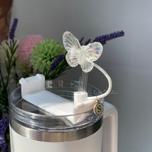 Puede incluir: Un vaso t&eacute;rmico de acero inoxidable blanco con una tapa transparente y un soporte de pajita de pl&aacute;stico blanco. La pajita tiene un encanto de mariposa iridiscente transparente unido a la parte superior. La pajita est&aacute; asegurada a la tapa con una correa de pl&aacute;stico blanco. Un encanto dorado con la letra "S" est&aacute; unido a la correa.