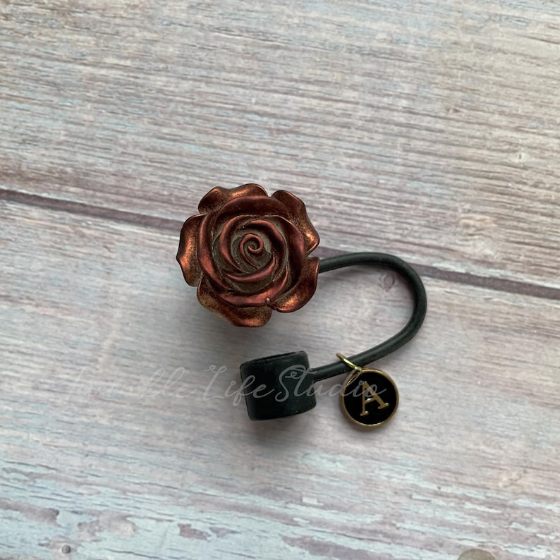 Pode incluir: Uma fivela de cabelo em forma de rosa cor de cobre com uma faixa el&aacute;stica preta e um berloque preto com a letra "A" em dourado. O acess&oacute;rio de cabelo est&aacute; sobre uma superf&iacute;cie clara com gr&atilde;o de madeira.