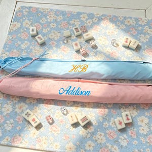 May include: Two long, cylindrical fabric cases in light blue and light pink, featuring drawstring closures. The blue case has gold embroidered initials HB, and the pink case has the name Addison embroidered in blue. Mahjong tiles and dice are scattered.