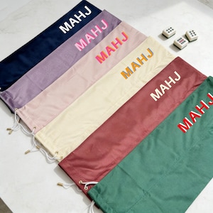 May include: Six rectangular fabric drawstring bags in navy, lavender, light pink, cream, maroon, and green. Each bag features the text MAHJ printed in a different colour. Three small white rectangular game tiles are positioned near the bags.