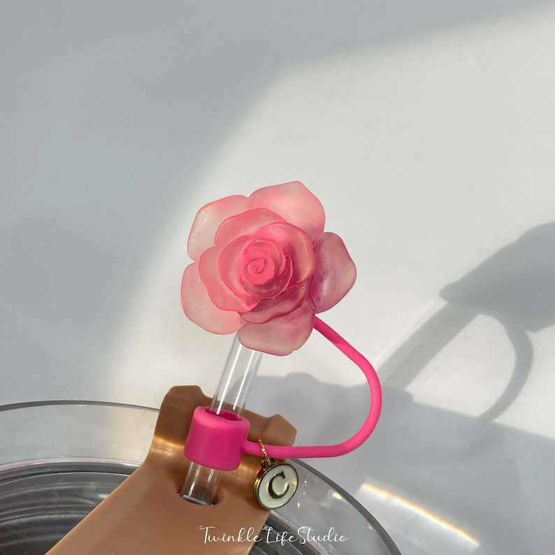 May include: A pink rose-shaped straw topper with a pink silicone straw holder and a gold charm with the letter "C" on it.