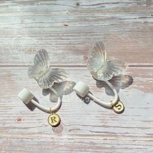 Puede incluir: Dos ganchos blancos en forma de mariposa con dijes dorados. Los dijes son redondos y tienen las letras "R" y "S" en ellos.