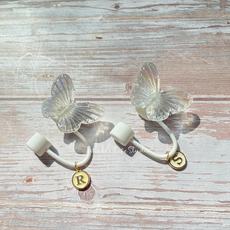 Puede incluir: Dos ganchos blancos en forma de mariposa con dijes dorados. Los dijes son redondos y tienen las letras "R" y "S" en ellos.