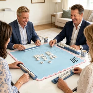 Pode incluir: Um tapete de mahjong quadrado azul-claro com uma borda rosa recortada e o monograma BC em preto. O tapete apresenta peças de mahjong brancas dispostas no centro e em quatro suportes azuis posicionados em cada lado da superfície de jogo.