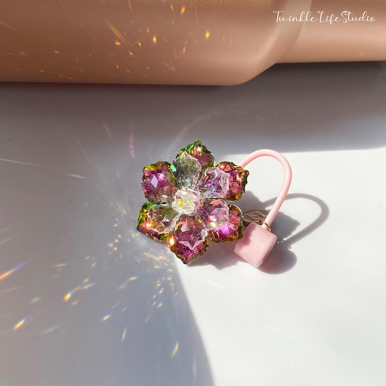 May include: A pink hair tie with a flower-shaped charm made of iridescent glass beads. The flower charm has a pink, green, and clear color scheme.