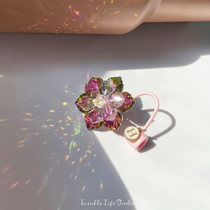 May include: A pink and green crystal flower charm with a pink elastic band and a gold charm with the letter "H" on it.