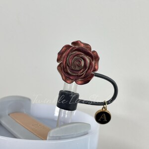 Pode incluir: Um tamp&atilde;o de garrafa decorativo com um design de rosa bord&ocirc;. Possui uma al&ccedil;a preta e um pingente dourado com a letra "A". O tamp&atilde;o est&aacute; preso a um tubo de vidro transparente.