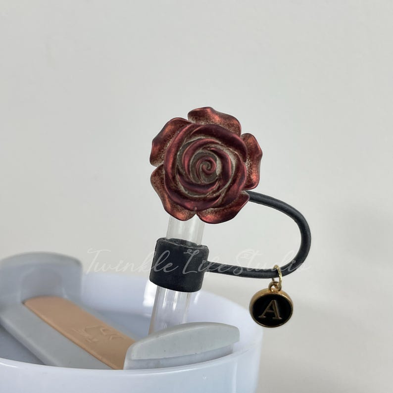 Pode incluir: Um tamp&atilde;o de garrafa decorativo com um design de rosa bord&ocirc;. Possui uma al&ccedil;a preta e um pingente dourado com a letra "A". O tamp&atilde;o est&aacute; preso a um tubo de vidro transparente.
