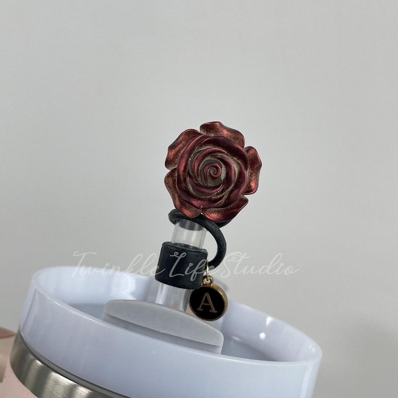 Pode incluir: Um tamp&atilde;o de garrafa decorativo com uma rosa vermelha met&aacute;lica detalhada. O tamp&atilde;o tem uma veda&ccedil;&atilde;o de borracha preta e um pingente dourado com a letra "A". O tamp&atilde;o &eacute; colocado em um copo branco.