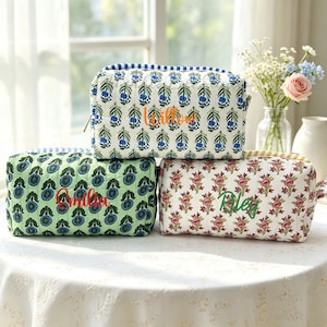 Könnte beinhalten: Drei rechteckige, gesteppte Stofftaschen mit Reißverschluss. Die obere Tasche hat ein blau-grünes Blumenmuster mit dem Namen Willow in Orange. Die untere linke Tasche ist hellgrün mit blauen Blumen und dem Namen Emilia in Rot.