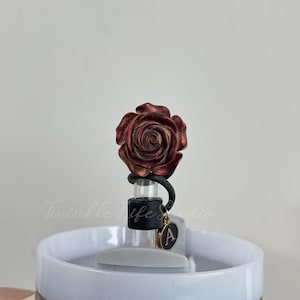 Pode incluir: Um tamp&atilde;o de garrafa decorativo com uma rosa met&aacute;lica detalhada em tons de vermelho e bronze. O tamp&atilde;o tem uma veda&ccedil;&atilde;o de borracha preta e um berloque dourado com a letra "A".