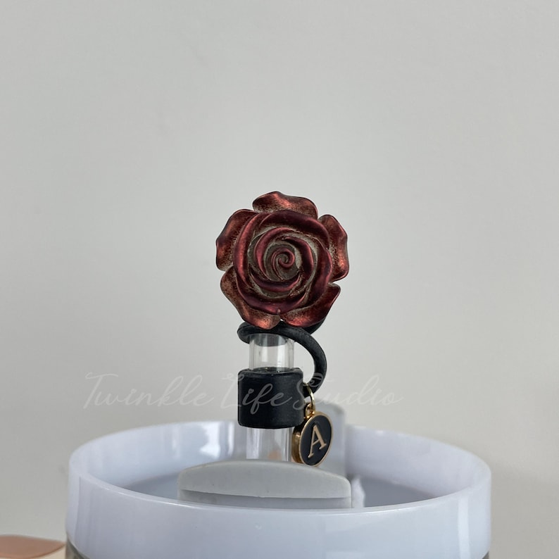 Pode incluir: Um tamp&atilde;o de garrafa decorativo com uma rosa met&aacute;lica detalhada em tons de vermelho e bronze. O tamp&atilde;o tem uma veda&ccedil;&atilde;o de borracha preta e um berloque dourado com a letra "A".