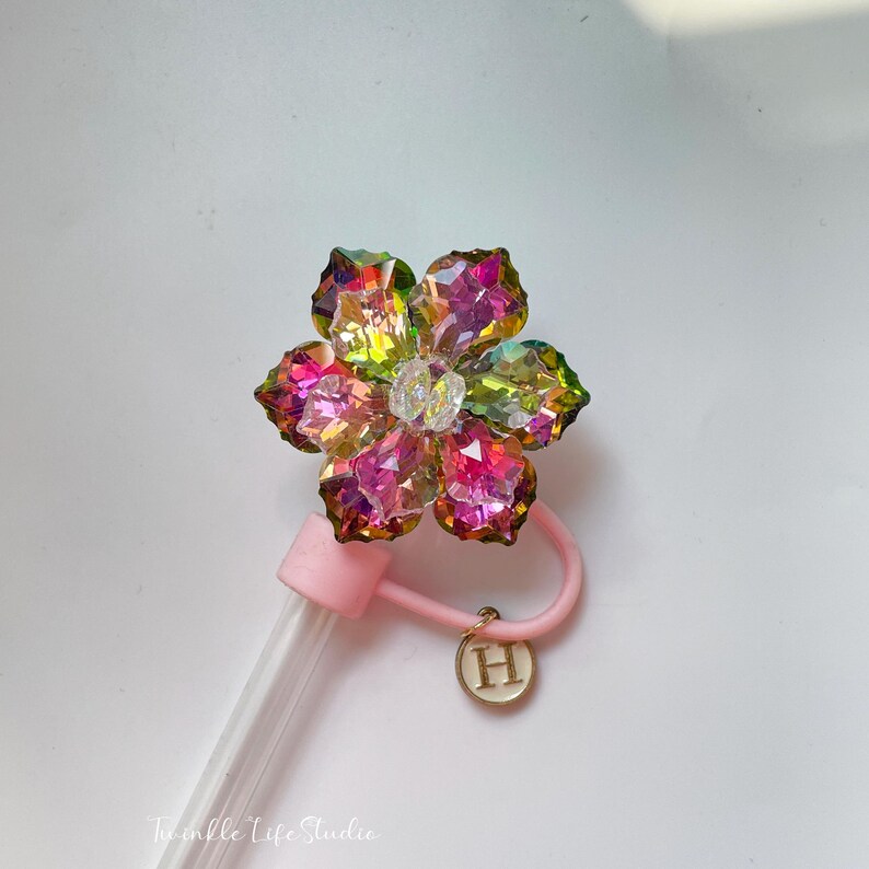 May include: A pink and clear glass bottle with a flower-shaped stopper made of iridescent crystals. The stopper is attached to a pink plastic handle with a gold charm that says "H".