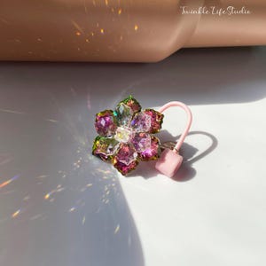 May include: A pink and green flower-shaped hair tie with iridescent crystals. The hair tie is made of clear elastic and has a pink rubber stopper.