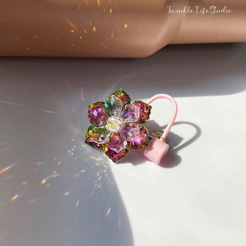 May include: A pink and green flower-shaped hair tie with iridescent crystals. The hair tie is made of clear elastic and has a pink rubber stopper.
