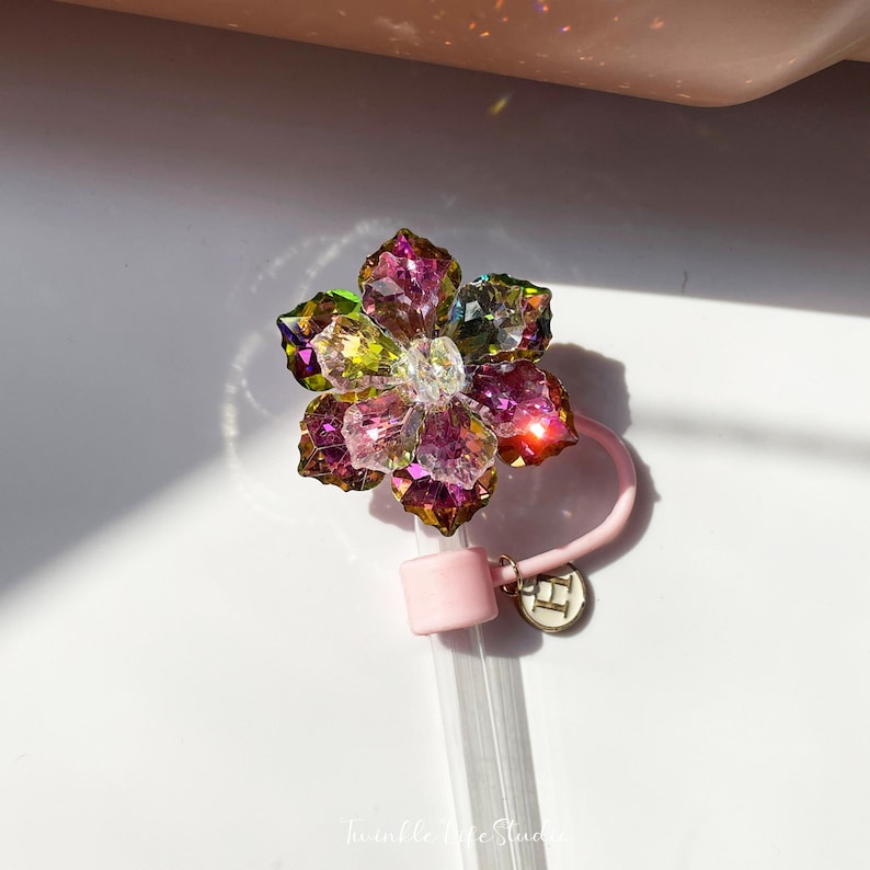 May include: A pink and clear glass straw with a flower-shaped charm made of iridescent crystals. The charm is attached to the straw with a pink silicone ring and a gold charm with the letter "H" on it.
