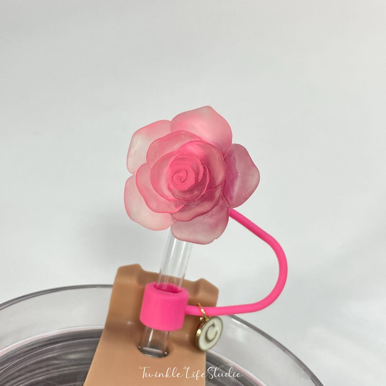 May include: A pink rose-shaped straw topper with a clear plastic straw and a pink silicone band. The straw topper is attached to a clear glass cup with a brown paper label that says "Twinkle Life Studio".