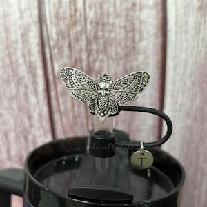 May include: A silver-colored, decorative butterfly bottle stopper with a skull design. The stopper is attached to a black lid with a small, round charm that has the letter "T" on it. The words "Twinkle Life Studio" are printed on the bottle.