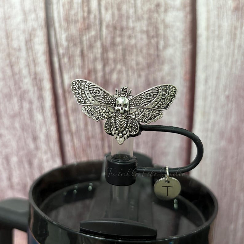 May include: A silver-colored, decorative butterfly bottle stopper with a skull design. The stopper is attached to a black lid with a small, round charm that has the letter "T" on it. The words "Twinkle Life Studio" are printed on the bottle.