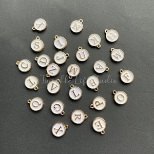 Puede incluir: Una colecci&oacute;n de 26 abalorios de metal blanco con detalles dorados, cada uno con una letra diferente del alfabeto. Los abalorios est&aacute;n esparcidos sobre una superficie gris oscura.