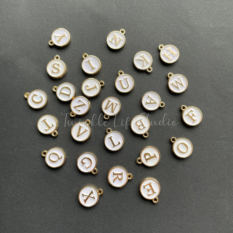 Puede incluir: Una colecci&oacute;n de 26 abalorios de metal blanco con detalles dorados, cada uno con una letra diferente del alfabeto. Los abalorios est&aacute;n esparcidos sobre una superficie gris oscura.