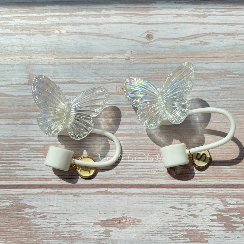 Puede incluir: Dos lazos para cortinas de mariposa blanca con alas iridiscentes y abalorios de letras doradas. Los abalorios est&aacute;n etiquetados con "S" y "T".