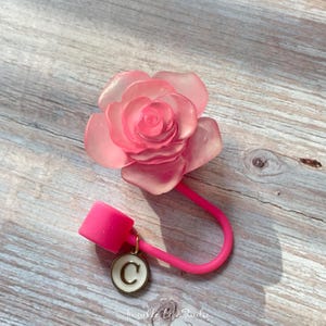 Könnte beinhalten: Ein rosa Silikon-Handyring mit einem durchsichtigen rosa Rosen-Anhänger und einem goldenen Anhänger mit dem Buchstaben C.