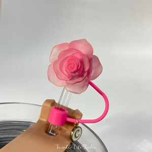 May include: A pink rose-shaped straw topper with a clear plastic straw and a pink silicone bendable straw holder. The straw holder is attached to a clear glass with a gold charm.