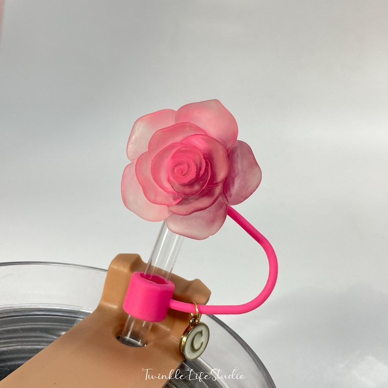May include: A pink rose-shaped straw topper with a clear plastic straw and a pink silicone bendable straw holder. The straw holder is attached to a clear glass with a gold charm.