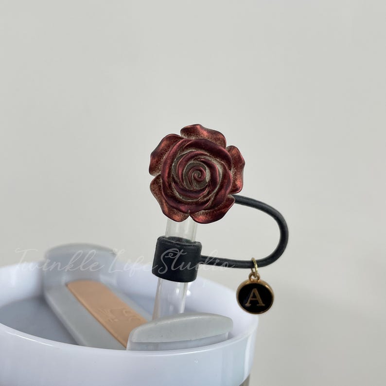 Pode incluir: Uma tampa decorativa para garrafa de &aacute;gua com um design de rosa bord&ocirc;. A tampa tem uma al&ccedil;a preta e um pequeno pingente dourado com a letra "A". A garrafa &eacute; branca com um gradiente azul claro.