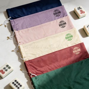 May include: A collection of six rectangular fabric drawstring pouches in navy, lavender, light pink, cream, maroon, and dark green. Each pouch features a circular stamp that reads In My Mahjong Era. Three mahjong tiles are placed near the pouches.