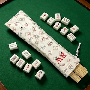 May include: A cream-coloured fabric storage bag featuring a repeating pattern of mahjong tile symbols and the red embroidered letters RW. The bag contains wooden tiles and is surrounded by several white mahjong tiles with red and black markings.