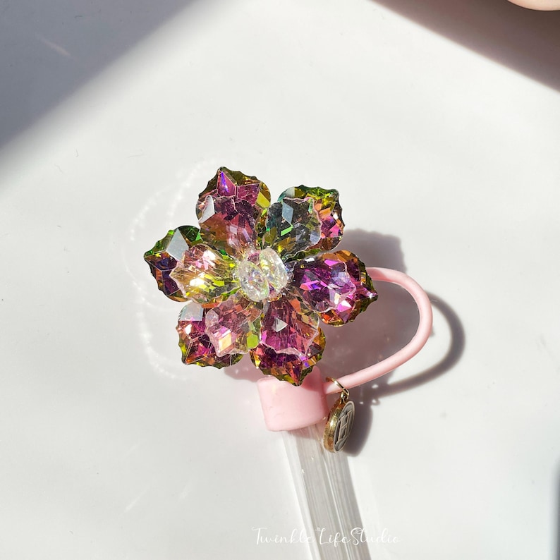 May include: A pink and clear plastic straw with a iridescent flower charm. The flower charm is made of faceted glass and has a pink, green, and purple color scheme.